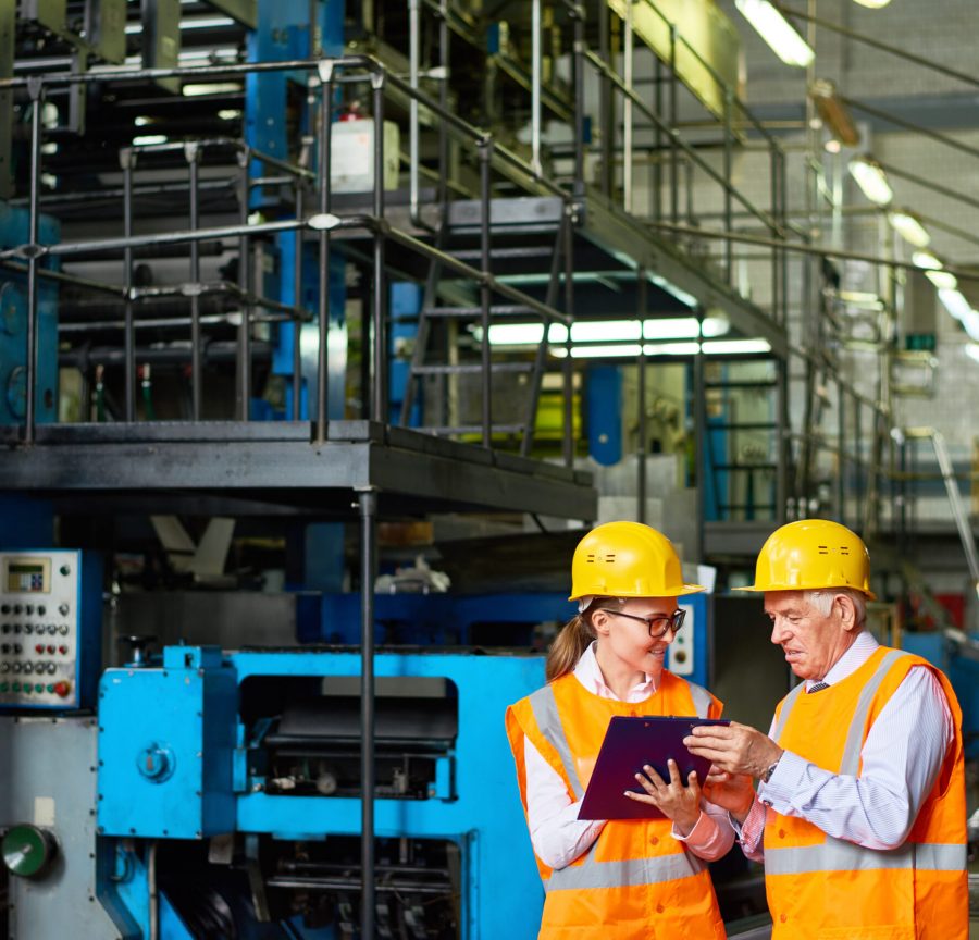 Portrait o senior factory worker discussing something with young woman in  modern workshop,  both wearing hardhats and reflective jackets